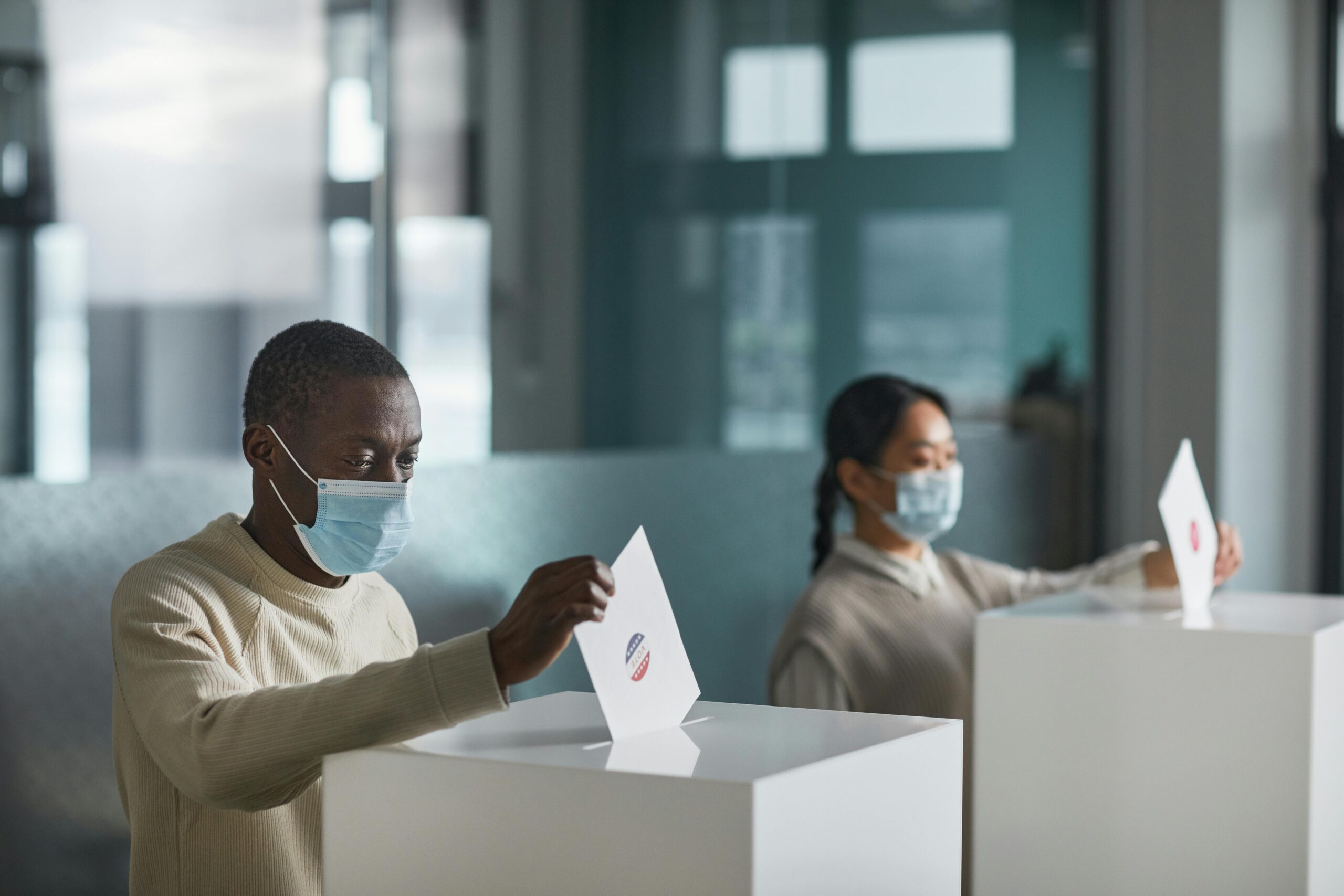 People wearing face masks cast ballots in a public indoor polling station with social distancing.